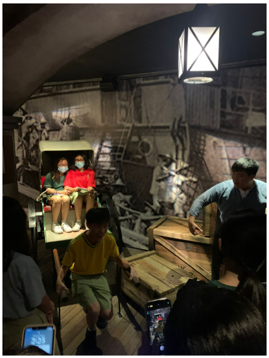 Two masked girls sit in a vintage carriage at a museum exhibit while children and adults explore around them.